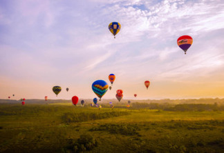 a imagem contempla um horizonte com céu azul alaranjado e vários balões de balonismo no céu, eles são bem coloridos e estão acima de um vasto campo verde.