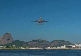 a imagem mostra um avião decolando do Aeroporto Santos Dumont, no Rio de Janeiro, com a Baía de Guanabara em frente e os morros da Urca e Pão de Açúcar ao fundo.