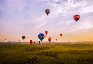 a imagem contempla um horizonte com céu azul alaranjado e vários balões de balonismo no céu, eles são bem coloridos e estão acima de um vasto campo verde.