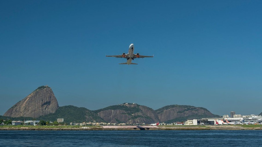 a imagem mostra um avião decolando do Aeroporto Santos Dumont, no Rio de Janeiro, com a Baía de Guanabara em frente e os morros da Urca e Pão de Açúcar ao fundo.