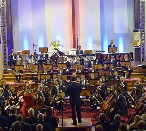 Em um palco, músicos de uma orquestra com um maestro no meio e parte do público na base da foto.