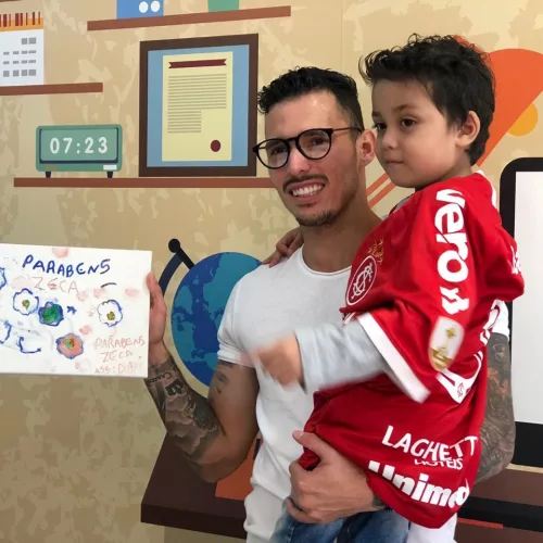 Jogador do Inter com menino no colo, vestindo camiseta vermelha do Inter. Na mão direita um desenho feito pelo menino dizendo parabéns