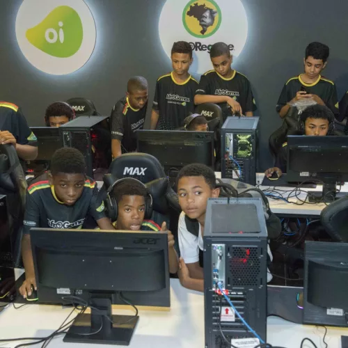 Em frente a computadores, jovens e crianças do projeto jogam durante a aula. Os meninos, que são maioria, são negros. No fundo, uma menina também participa da atividade.