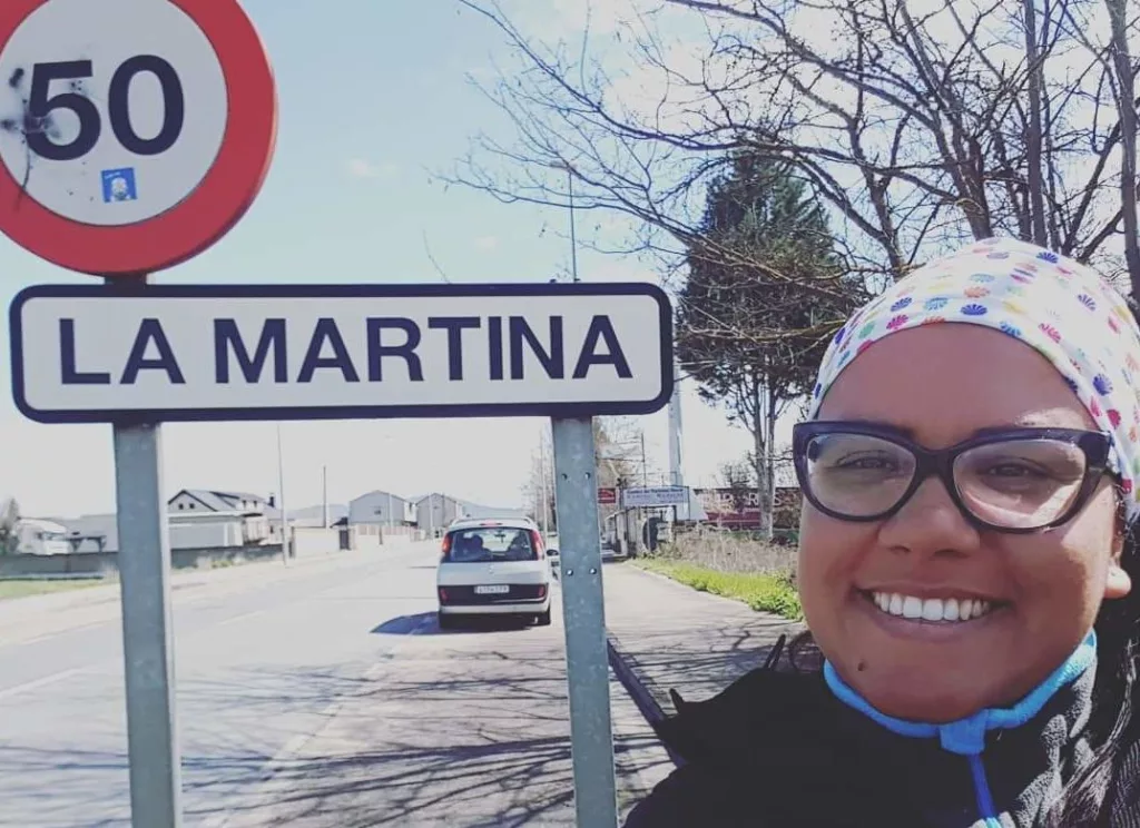 Um mulher, usando toca e óculos, ao lado de duas placas, uma indicativa da velocidade de 50km, e outra com a inscrição La Martina.
