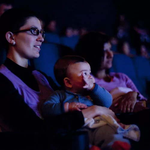 Mulheres com crianças no colo comem pipoca e olham para a tela do cinema em uma sala parcialmente iluminada.