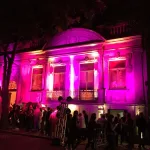 Fachada de prédio histórico iluminada em rosa com várias pessoas em fila