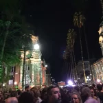 Nas pontas, dois prédios iluminados com figuras. Ao centro, centenas de pessoas reunidas em uma praça.