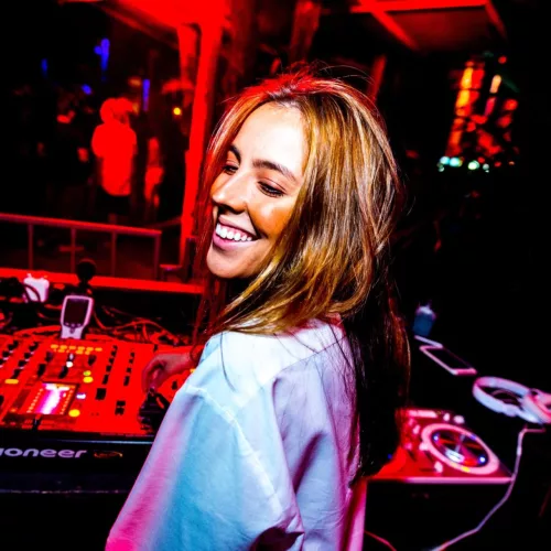 A DJ Mari Hermel, em frente a uma mesa de som, em um ambiente de luz avermelhada.