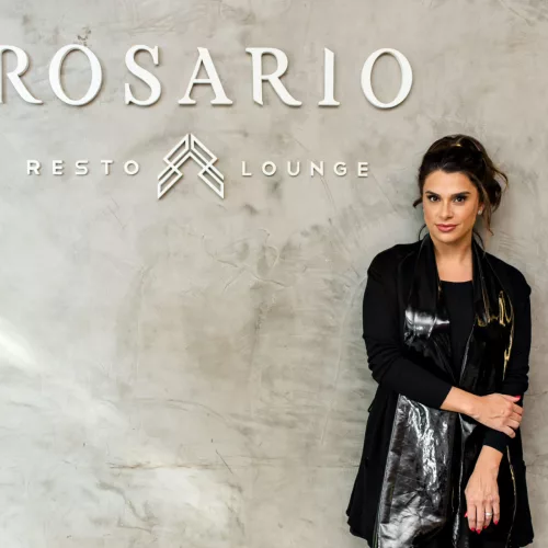 Mulher vestida de preto, encostada na parede, onde se lê Rosario Resto Lounge
