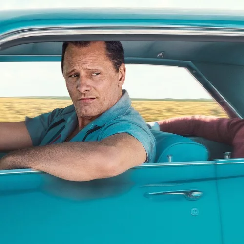 Um homem branco no lugar do motorista, e outro, negro, no banco de trás de um carro cor verde azulado ou azul esverdeado.