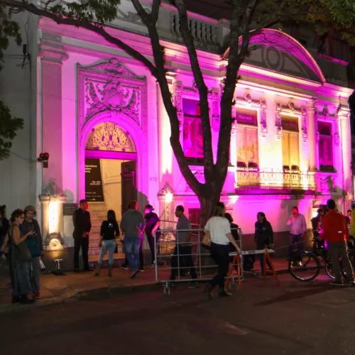 À noite, diversas pessoas se concentram em frente a um prédio antigo, iluminado, em tons de rosa.