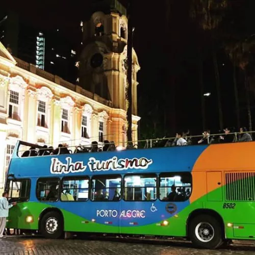 Ônibus turístico colorido, com as inscrições Linha Turismo e Porto Alegre. Ao fundo, iluminado, um prédio histórico, o Museu de Arte do RS, o MARGS.
