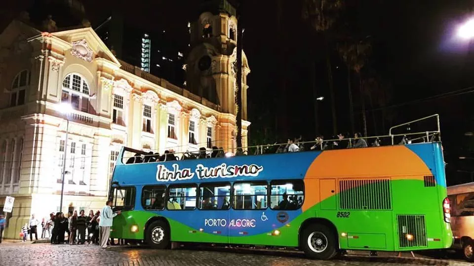 Ônibus turístico colorido, com as inscrições Linha Turismo e Porto Alegre. Ao fundo, iluminado, um prédio histórico, o Museu de Arte do RS, o MARGS.