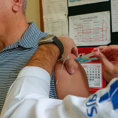 Um homem com jaleco aplica vacina em um homem aparentando mais de 50 anos. No jaleco lê-se SUS.