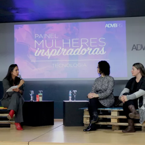 Três mulheres, sentadas, uma delas com um microfone, falam em um palco, onde atrás está projetado "Painel Mulheres inspiradoras - Tecnologia" e ADVBRS.