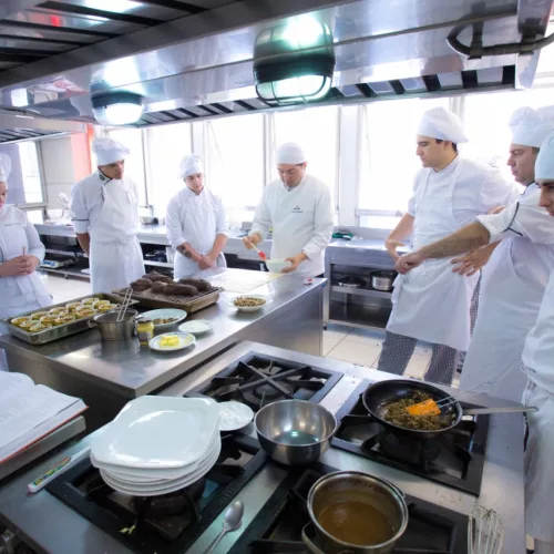 Diversas pessoas, ao redor de uma cozinha equipada, com aventais e chapéus próprios para a prática culinária.