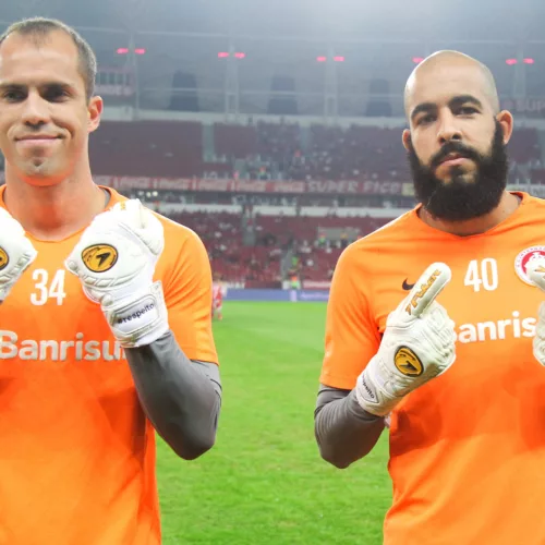 Dois goleiros, com fardamento em cor laranja, dentro do estádio, mostram as luvas com a hashtag da campanha.