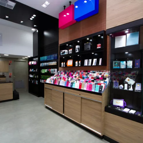 Um espaço com móvel tipo estante mostruário de madeira contendo diversas opções de capas coloridas e acessórios para celular. Ao fundo lê-se o nome da loja - I Like Mobis.