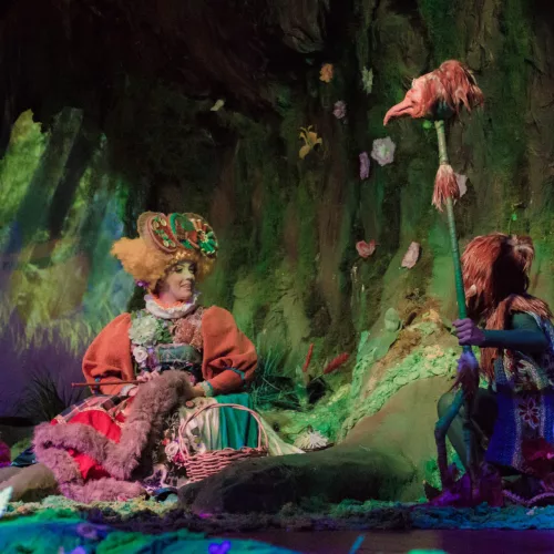 Em um cenário predominantemente verde, vê-se quatro pessoas com figurino colorido, mangas bufantes, vestidos, grandes chapéus.