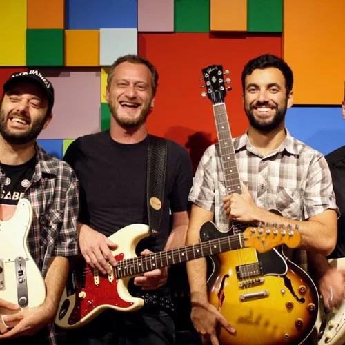 Quatro homens com guitarras sorriem em frente a uma parede com quadrados coloridos.