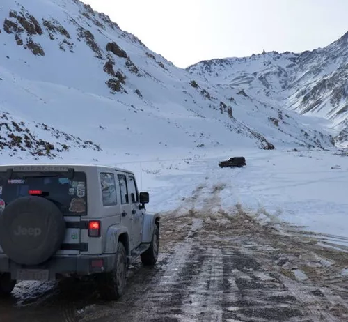 Um veículo Jeep percorre um estrada e meio à neve, enquanto outro está mais à frente. Ao fundo vê-se montanhas nevadas.