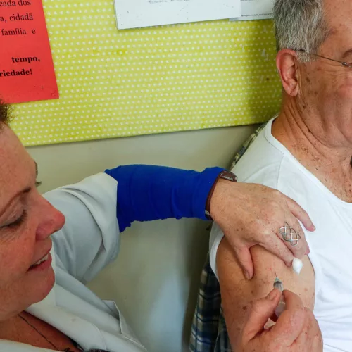 Um mulher aplica uma injeção no braço de um idoso, que sorri.