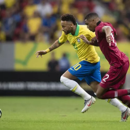 Jogador da Seleção Brasileira, de camiseta amarela 10 e calção azul, tenta se desvencilhar do jogador da seleção do Catar, camisa 2, vestindo uniforme cor vinho.