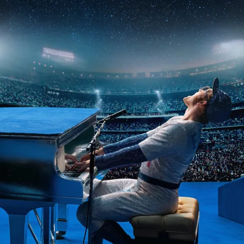 Um homem toca ao piano em um palco, jogando o corpo para trás. Ao fundo vê-se um estádio lotado. A iluminação é predominantemente azul.