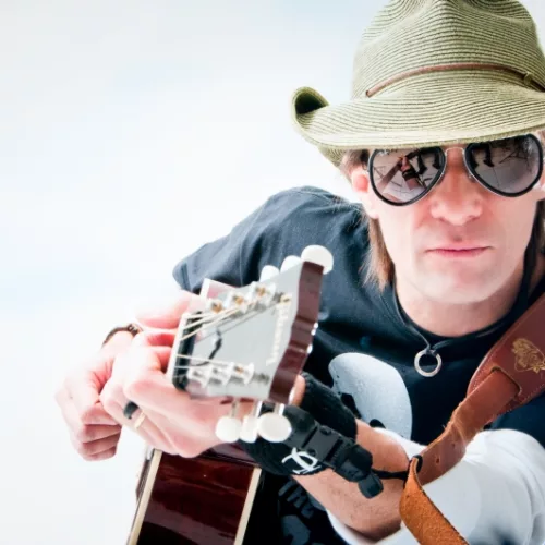 Um homem com chapéu e óculos escuros empunha um violão.
