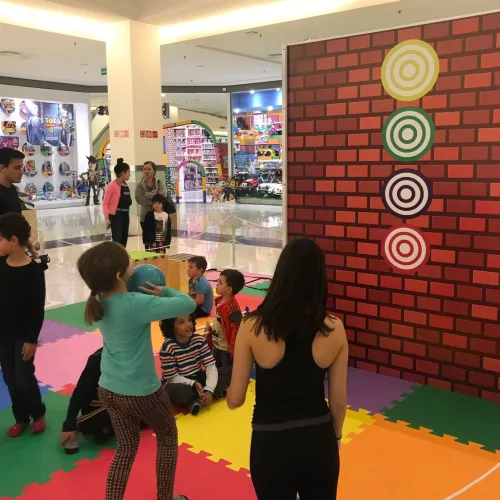 Crianças brincam de tiro ao alvo com uma bola em um espaço colorido.