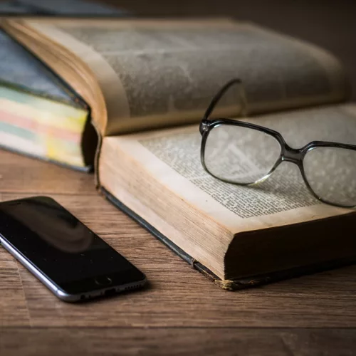 Livros abertos com um óculos em cima e um celular ao lado.
