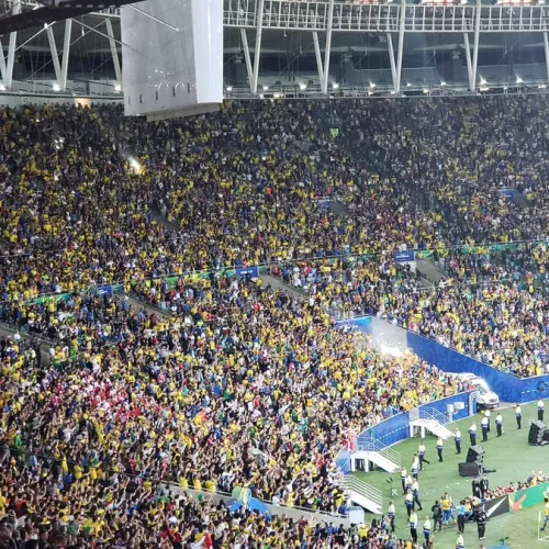 Parte de um estádio com torcedores do Brasil em jogo da Seleção. No canto inferior direito vê-se alguns jogadores brasileiros.