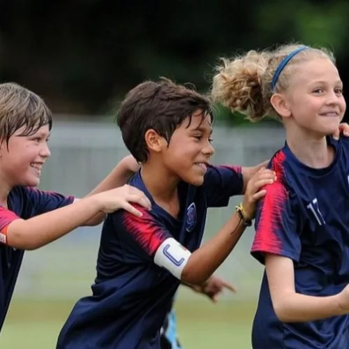 Uma menina loira corre com dois meninos atrás, uniformizados e em um campo de futebol, aparentemente comemorando após um gol