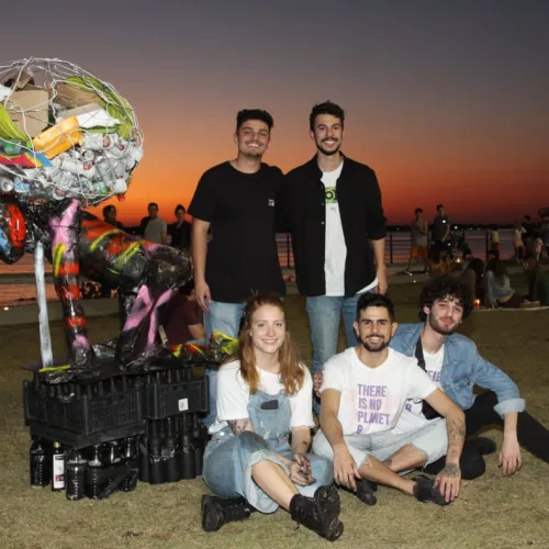 Cinco jovens, quatro rapazes e uma garota, três sentados e dois em pé, ao lado de materiais reciclados. Eles estão na orla do Guaíba, onde atrás o sol se põe.
