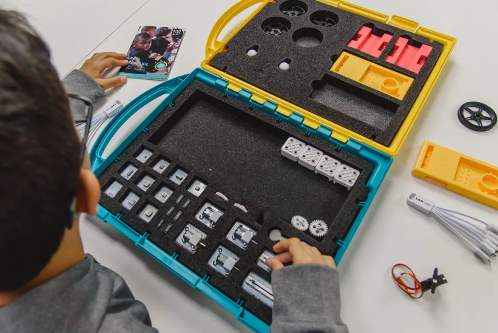 Uma criança mexe em uma espécie de maleta colorida com várias peças, como dados.