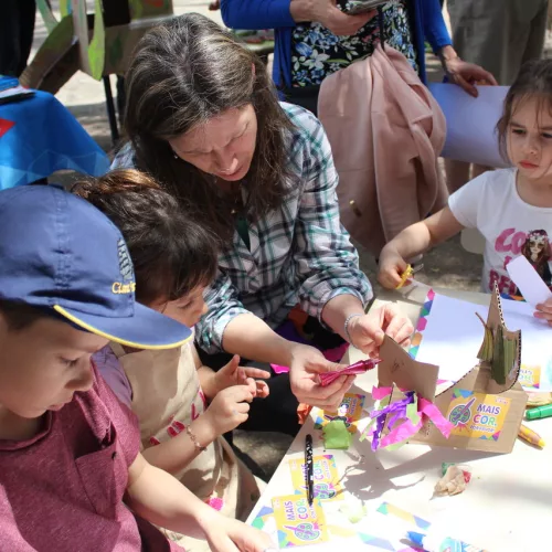 uma adulta e três crianças brincam em um mesa com lápis de cor, cola, papel e muitas cores. Atrás mais pessoas e lápis coloridos.