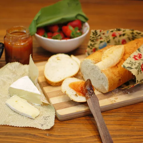 Embalagens podem ser usadas para embalar alimentos diversos, como pães, na foto em cima de uma tábua de madeira, queijos ou frutas. Na foto, pão, queijo, geleia e um pote de morangos.