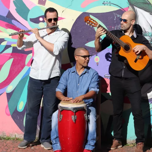 Fiesta latina: três homens, um com violão, outro com flauta, ambos em pé, atrás um muro colorido. Sentado, um homem na percussão.