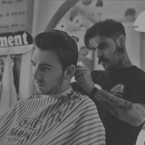 Um homem corta o cabelo de outro em um barbearia. Foto em preto e branco. O homem que tem o cabelo cortado usa um avental listrado.