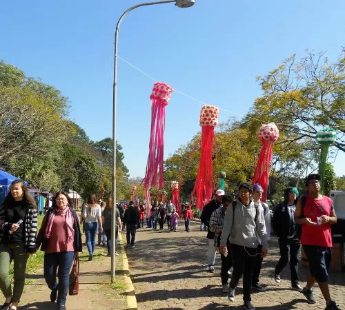 8ª edição do Festival do Japão. Pessoas caminham em uma área ao ar livre, com árvores ao redor, num dia de céu azul. Ao fundo, alegorias.