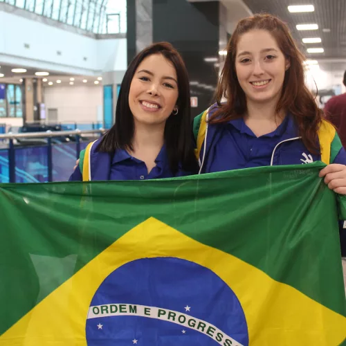 WorldSkills . Duas mulheres jovens vestindo azul empunham a bandeira do Brasil no aeroporto de Porto Alegre.
