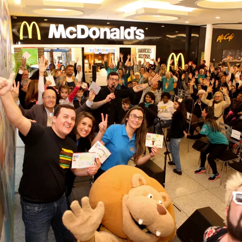 McDia Feliz 2019, diversas pessoas fazem sinal de positivo e vitória em frente a um palco montado em frente a um McDonalds. O mascote do ICI também está presente.