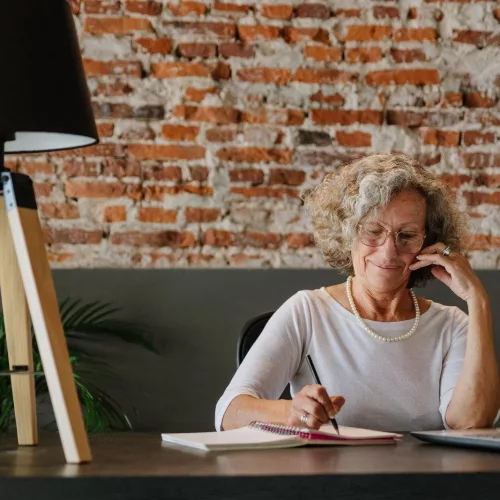 Uma mulher, aparentemente em torno dos 50 anos, está na frente de um notebook, enquanto faz anotações em um caderno, em uma mesa, com um abajur ao seu lado. Atrás, uma parede com tijolos à vista.