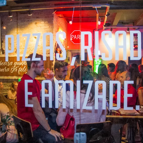 Vista de fora para dentro da janela do Na Vida Real Bar. Lê-se Pizzas, risadas e amizade em adesivo na janela.