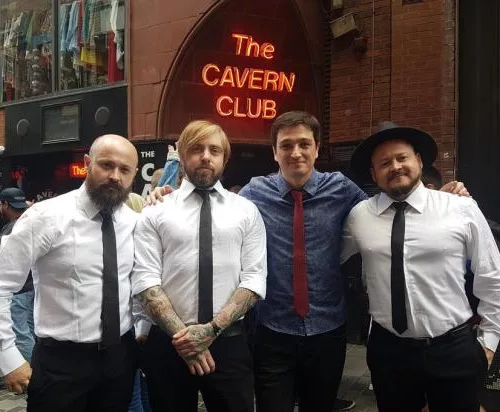The Beatles no Acordeon : Quatro homens de gravata, três deles com camisa branca e gravata preta e um com camisa azul e gravata vermelha em frente ao bar The Cavern Club.