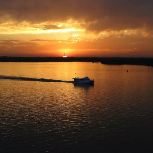 Pôr do sol no Guaíba. Um barco atravessa o curso de água sob um céu de sol entre muitas nuvens.
