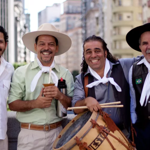 Os Fagundes: quatro homens vestindo roupas gaúchas, um deles carrega instrumento de percussão.
