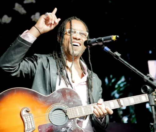 Serginho Moah. Um homem negro, de óculos e tranças, segura um violão em frente a um microfone, enquanto sorri e aponta para cima.