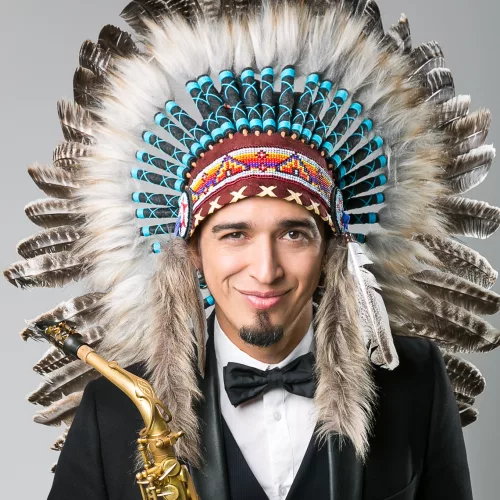 Vini Netto. Um homem usando smoking e um cocar de índio estilo americano segura um saxofone