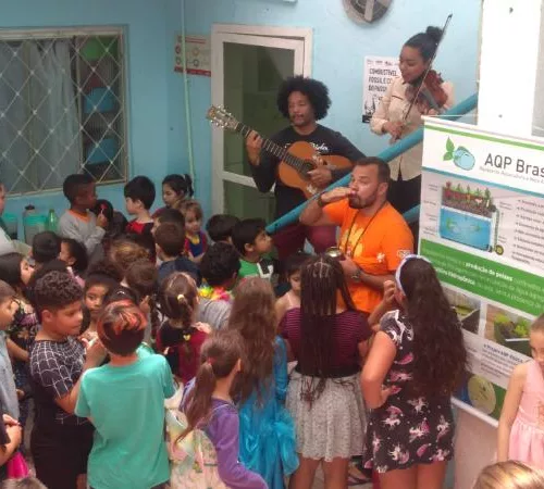 Sistema de energia solar em uma creche. Diversas crianças assistem a adultos que tocam música com violão.